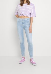 Camiseta corta de teñido anudado en morado, jeans skinny de color azul claro y zapatillas de ante rosas. Las zapatillas cuentan con suelas blancas y un diseño de cordones.