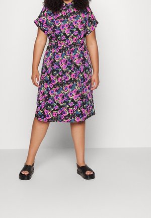 Femme portant une robe noire longueur genoux avec un imprimé floral rose et violet, manches courtes, et des sandales plateformes noires, debout sur un sol gris.