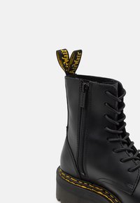 Stivale alla caviglia in pelle nera con un chunky platform, chiusura con laccifrontale, zip laterale e dettagli di cucitura gialla sulla suola.