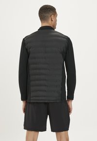 Schwarzige Weste mit gepolstertem Design und glatter Textur. Ein schwarzes Langarmshirt und passende Shorts runden das Outfit ab.