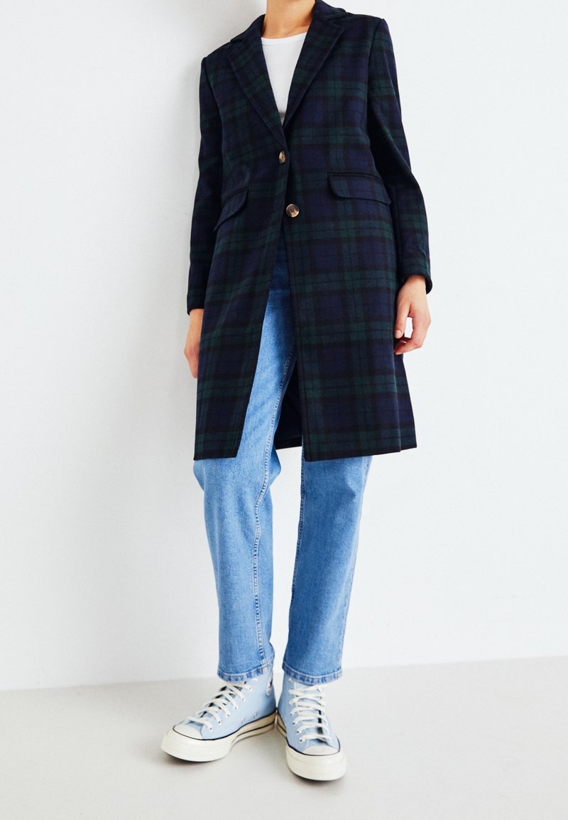 Dlouhý károvaný kabát v námořnické modři a zelené barvě, s jedním knoflíkem, špičatými klopami, bočními kapsami, v kombinaci se světle modrými džínami a teniskami.
