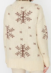 Cremfarbener Strickpullover mit braunen Schneeflockenmustern, geripptem Saum und lockerer Passform mit sichtbaren Nahtdetails.