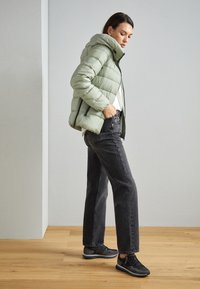 Leichte, grüne Pufferjacke mit Kapuze, Reißverschluss vorne und seitlichen Taschen, kombiniert mit hochgeschnittenen, dunkelgrauen Jeans und schwarzen Turnschuhen.