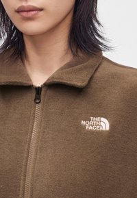 Personne aux cheveux mi-longs et foncés portant une veste polaire marron zippée avec le logo The North Face sur la poitrine.