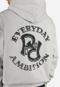 Grå hættetrøje med en stor sort grafisk tryk på ryggen med teksten "EVERYDAY AMBITION" og stiliseret 'R'. Blød tekstur.