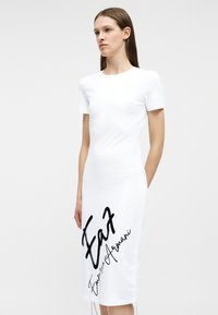 Γυναίκα που φοράει λευκό midi φόρεμα με κοντά μανίκια, με μαύρη γραφή "Emporio Armani" και "EA7" τυπωμένη στην μπροστινή αριστερή πλευρά.