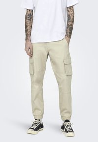 Beige cargobroeken hebben een losse pasvorm met elastische boorden, meerdere zijzakken en verstevigde knieën. Gepaard met zwarte sneakers.