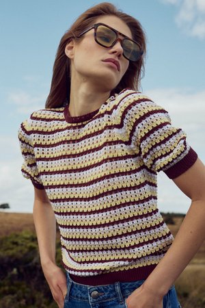 Femeie în aer liber purtând ochelari de soare mari cu rame de tip scoică țestoasă și un top tricotat cu mâneci scurte, în dungi, în culorile vișiniu, galben și alb.