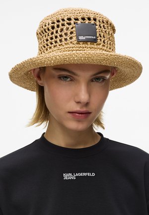 Νεαρός που φοράει μπεζ πλεγμένο ψάθινο καπέλο με μαύρο τετράγωνο λογότυπο και μαύρο πουκάμισο με λευκό κείμενο «Karl Lagerfeld Jeans».