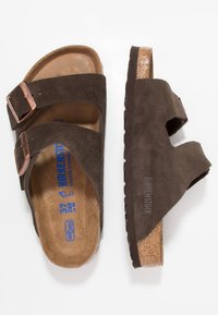 Sandales en daim marron avec deux strap ajustables, semelle en liège et semelle en caoutchouc texturé. Présente un logo Birkenstock sur le côté.