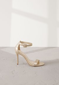 Sandale à talon haut métallisée dorée avec bride cheville et boucle, design à bout ouvert, texture lisse et talon aiguille fin.