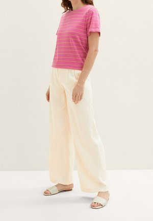 Kobieta ubrana w różowy, krótkim rękawem, pasiasty top, szerokie, kremowe spodnie i beżowe klapki, stojąca na jasnym tle.