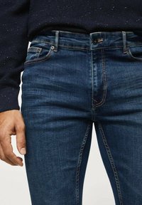 Jean en denim bleu foncé avec une coupe slim, présentant un délavage subtil et des surpiqûres oranges contrastantes. Comprend des poches avant et une fermeture à bouton en métal.