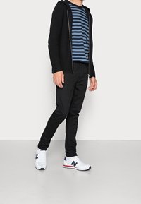 Svart hoodie med dragkedja, marinblå och blå randig t-shirt, svarta jeans, vita sneakers med röda och marinblå detaljer. Släta texturer, figursydd design.