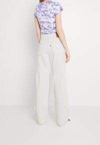 Jeans de pierna ancha en gris claro con bolsillos traseros cosidos y una etiqueta de cuero. Combinados con una camiseta de manga corta de tinte anudado morado.