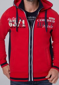 Rode zip-up hoodie met marineblauwe accenten, voorzien van geprinte luchtvaartgraphics, een grote capuchon en ribgebreide manchetten en zoom. Soepele textuur, casual pasvorm.