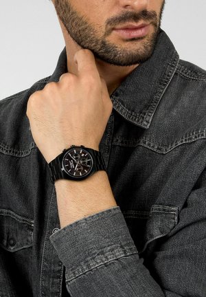 Orologio da polso nero con quadrante rotondo, quadrante nero texturizzato, dettagli in argento e bracciale metallico. Indossato su un polso con una giacca di jeans grigia.
