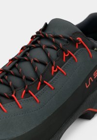 Zapatilla deportiva negra y gris oscuro con cordones negros acentuados en rojo y lazos para los cordones rojos, que presenta una parte superior de tela texturizada y una suela gruesa.