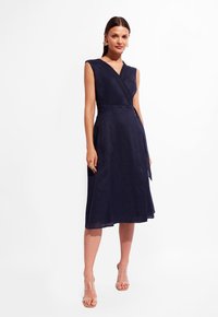 Granatowa lniana sukienka wrap z bezrękawnikowym krojem, fakturowanym wykończeniem, paskiem w talii oraz sylwetką w kształcie litery A, dopełniona przez przezroczyste sandały na obcasie.