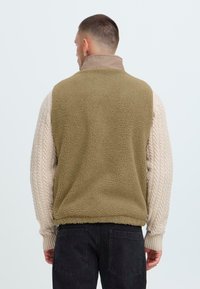 Fleece vest with olive green back, cream cable-knit sleeves, high collar, and textured fabric. Black trousers complete the look.