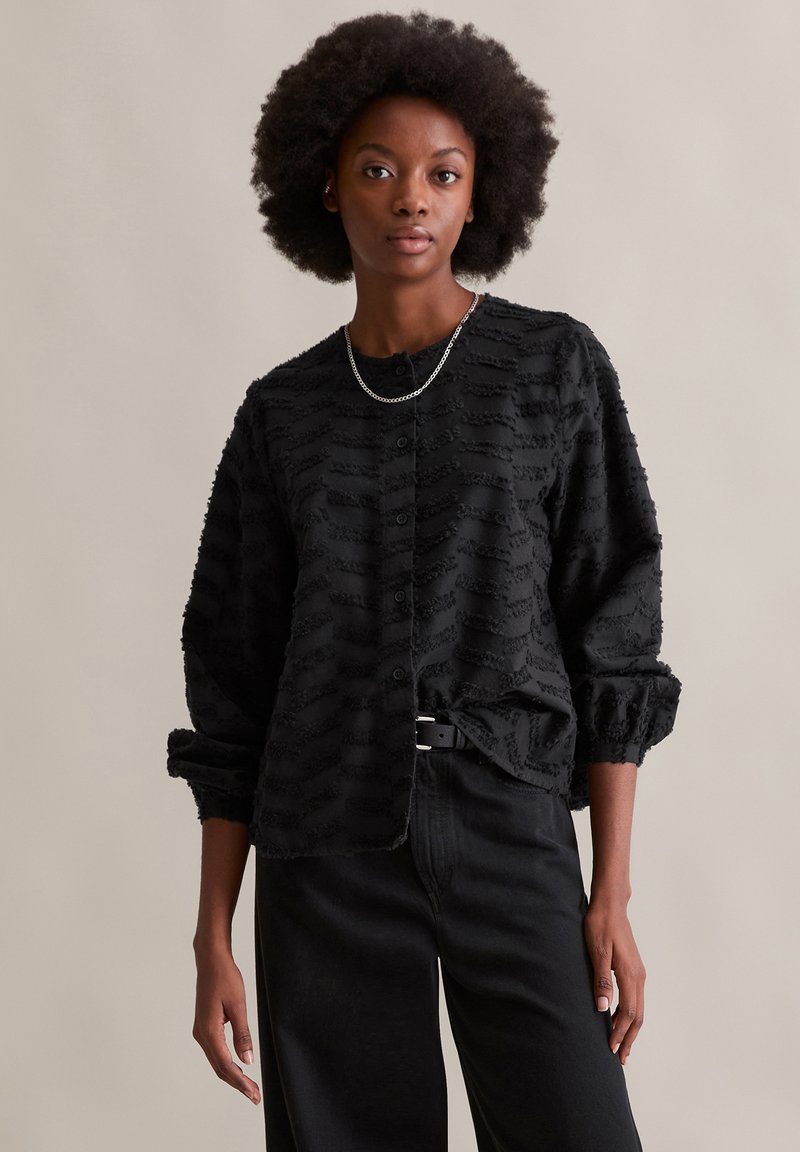 Schwarze Baumwollhemd mit strukturiertem Gewebe und gewellten Mustern, mit gerafften Ärmeln. Trägt man über hochgeschnittenen schwarzen Hosen. Silberne Kettenhalskette.