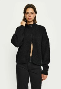 Schwarze gestrickte Strickjacke mit hohem Kragen, durchgehendem Reißverschluss und geripptem Saum und Ärmelbündchen. Weiche Textur und lässige Passform mit überschnittenen Schultern.
