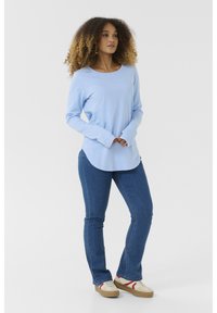 Helleblaue Langarmbluse mit abgerundetem Saum, kombiniert mit dunkelblauen Jeans und beigen Sneakers mit roten Akzenten.