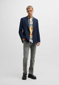 Blazer doppiopetto blu sul t-shirt grafica con una stampa colorata. Jeans grigi slim-fit e scarpe nere con suola spessa completano l'outfit.