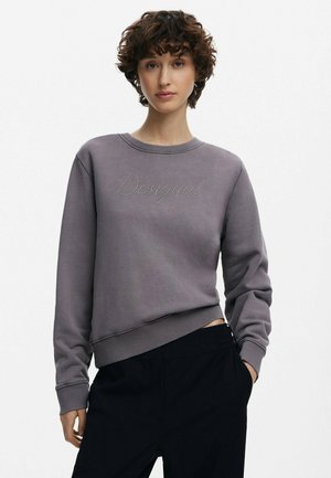Sudadera gris con cuello redondo, puños y dobladillo acanalados. Presenta "Desigual" en pedrería en la parte frontal. Combinada con pantalones oscuros.