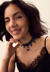 Black lace choker with bead accents, featuring intricate detailing. Model wearing a black lace-trimmed camisole with wavy hair and a gentle smile.