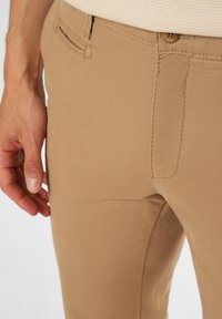 Pantalon beige avec une surface lisse et texturée. Présente une poche latérale et un détail de couture subtil, mettant en valeur une coupe ajustée.