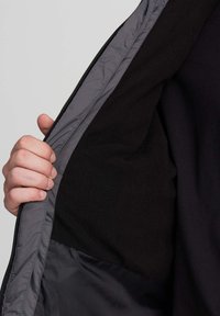 Una mano tiene aperto un giaccone grigio che mostra un rivestimento in pile nero e una tasca interna cucita su uno sfondo semplice.
