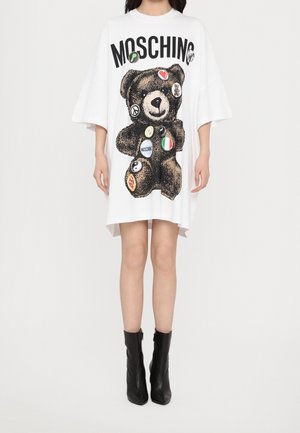 Femme portant une robe blanche oversize avec un grand ours en peluche et des logos imprimés sur le devant, associée à des bottines noires.