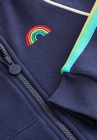 Granatowa kurtka zapinana na zamek z kolorową haftowaną tęczą i kontrastowym zielonym paskiem wzdłuż ramienia. Miękki, teksturowany materiał.