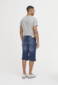 Camiseta gris de manga corta combinada con pantalones cortos Bermuda de mezclilla azul oscuro, que cuentan con bolsillos traseros y un ligero desvanecimiento. Zapatillas grises completan el look.