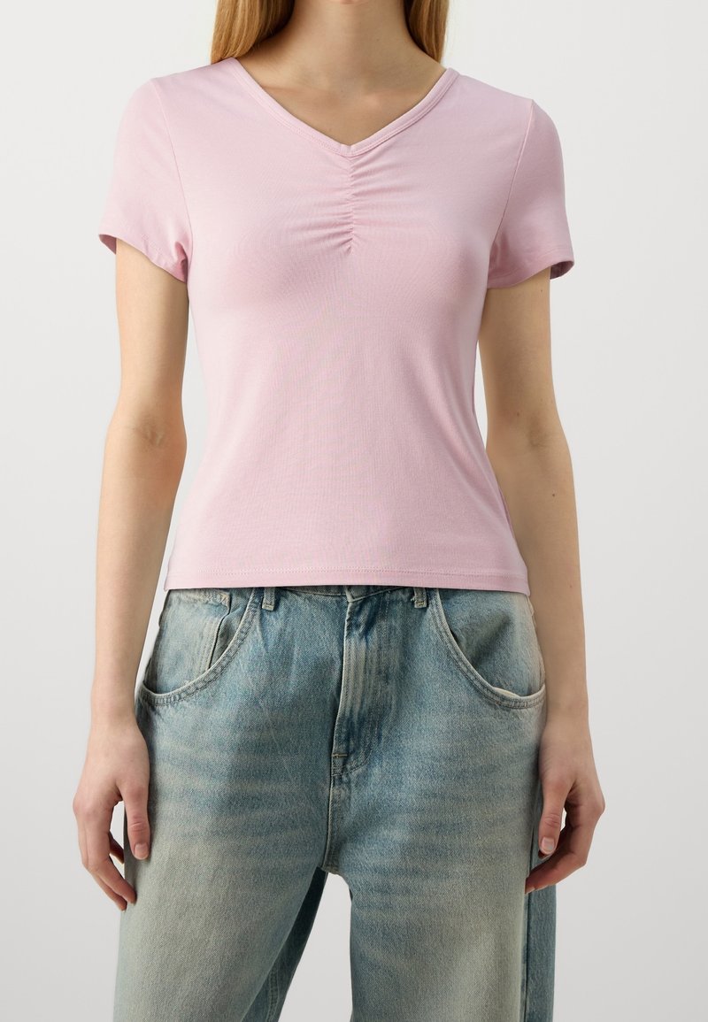 Femme portant un haut rose clair à manches courtes et col en V avec un détail froncé, associé à un jean bleu délavé ample avec des poches avant.