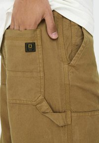 Brune canvas cargo bukser med flere lommer, synlig syning og et lille sort mærke. Struktureret stof, robust design.