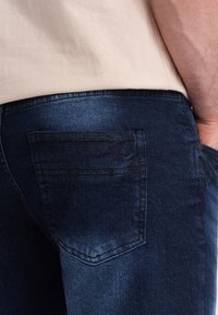 Jeans bleu foncé avec une poche arrière standard et des détails de couture. Texture en denim douce, légèrement délavée pour un aspect usé.