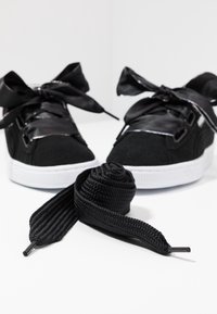 Paire de baskets noires avec des lacets en ruban de satin noués en nœuds, semelles blanches, et un lacet noir supplémentaire enroulé devant sur une surface blanche.