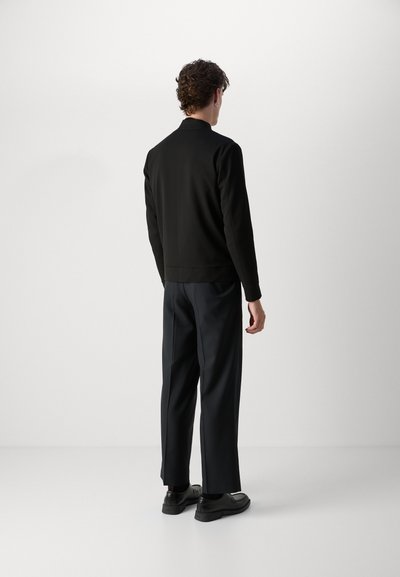 Pull noir à col montant, texture lisse et ourlet côtelé. Associé à un pantalon large et sombre et des chaussures noires.