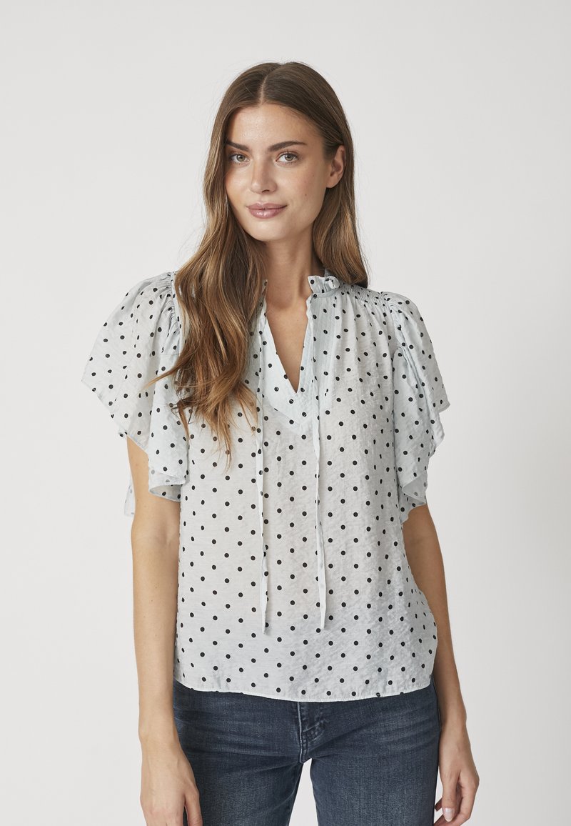 Blusa corta a maniche corte di colore azzurro chiaro con pois neri, scollatura a V e maniche arricciate. Il materiale sembra leggero e leggermente trasparente.
