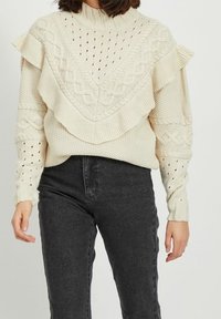 Pull en maille crème avec un motif texturé, doté d'un froufrou festonné le long du décolleté et de motifs en points ajourés sur les manches.
