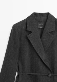 Grå koksgrå frakke med slagkrave, bælte og knappe detaljer. Fremstillet i tekstureret stof med skræddersyet pasform og elegant design.