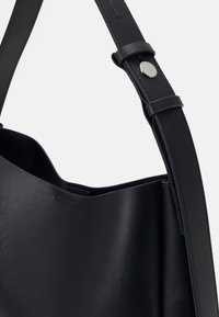 Sac à épaule en cuir noir avec une texture lisse, une large ouverture et une sangle plate dotée d'un fermoir à pression en argent pour l'ajustement.