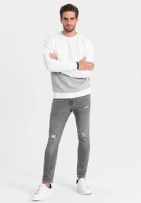Weiß und grau farblich abgesetzter Pullover mit Rundhalsausschnitt, kombiniert mit grauen, ausgebleichten Jeans und weißen Turnschuhen. Glatte Textur, lässige Passform.