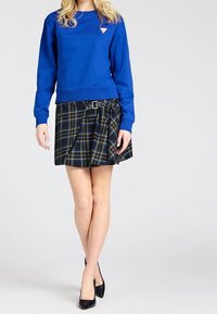 Mujer vistiendo un sudadera azul, una falda de cuadros plisada en negro y azul con cinturón, y tacones altos negros, de pie contra un fondo liso.