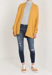 Senapsgul oversized stickad cardigan, grå högkragad topp, sliten mörk jeans och vita sneakers med bruna detaljer.