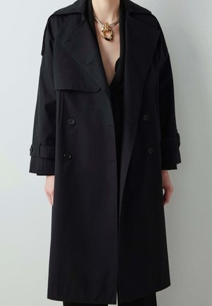 Person, die einen langen schwarzen doppelreihigen Trenchcoat mit breitem Revers und eine klobige goldene Kette mit großem Anhänger trägt.