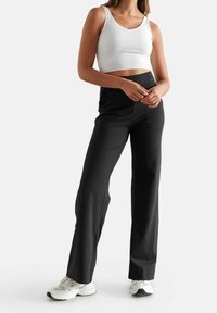 Pantalons noirs taille haute avec une texture lisse et des jambes larges, associés à un débardeur blanc ajusté et des chaussures de sport blanches.