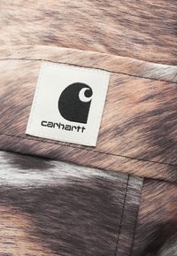 Carhartt-stof met een getextureerd, abstract patroon in tinten bruin en grijs, met een witte label waarop het Carhartt-logo staat.